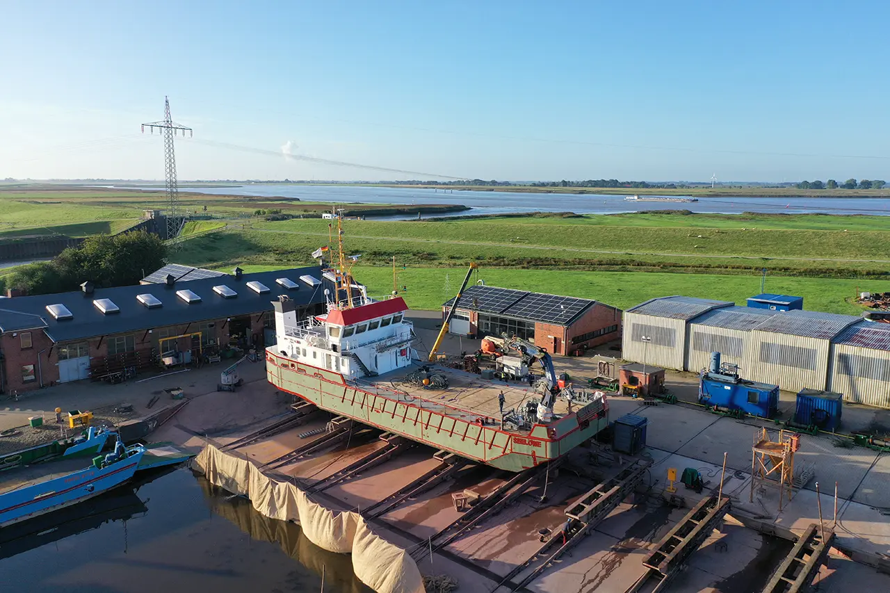 Luftaufnahme einer Werft an einem Fluss: Das Schiff „Seelöwe“ liegt mit grüner Grundierung auf der Helling, daneben Werkhallen und Marschwiesen.