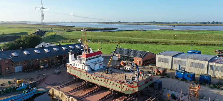Luftaufnahme einer Werft an einem Fluss: Das Schiff „Seelöwe“ liegt mit grüner Grundierung auf der Helling, daneben Werkhallen und Marschwiesen.