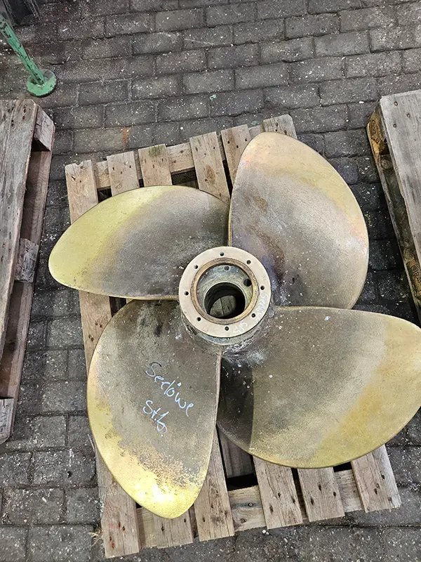 Alter, abgenutzter Vierblatt-Propeller aus Bronze auf einer Holzpalette, mit Kreide als „Seelöwe Stb“ beschriftet.