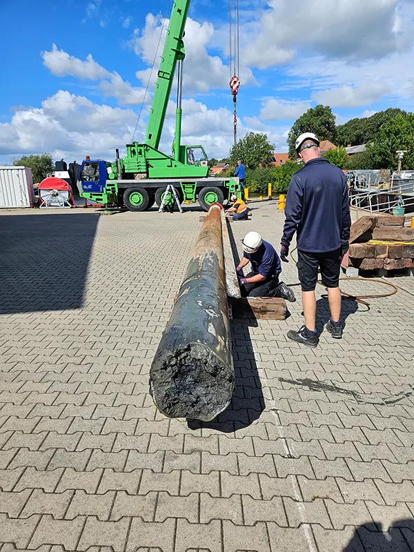 Ausgebaute Welle liegt auf Holzböcken am Boden, zwei Werftmitarbeiter mit Schutzhelm begutachten sie.