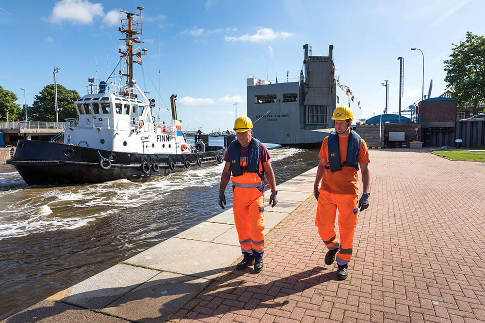 Festmacher im Hafen bei der Arbeit Zwei Festmacher laufen entlang der Schleusenkante, im Hintergrund ein Schlepperschiff