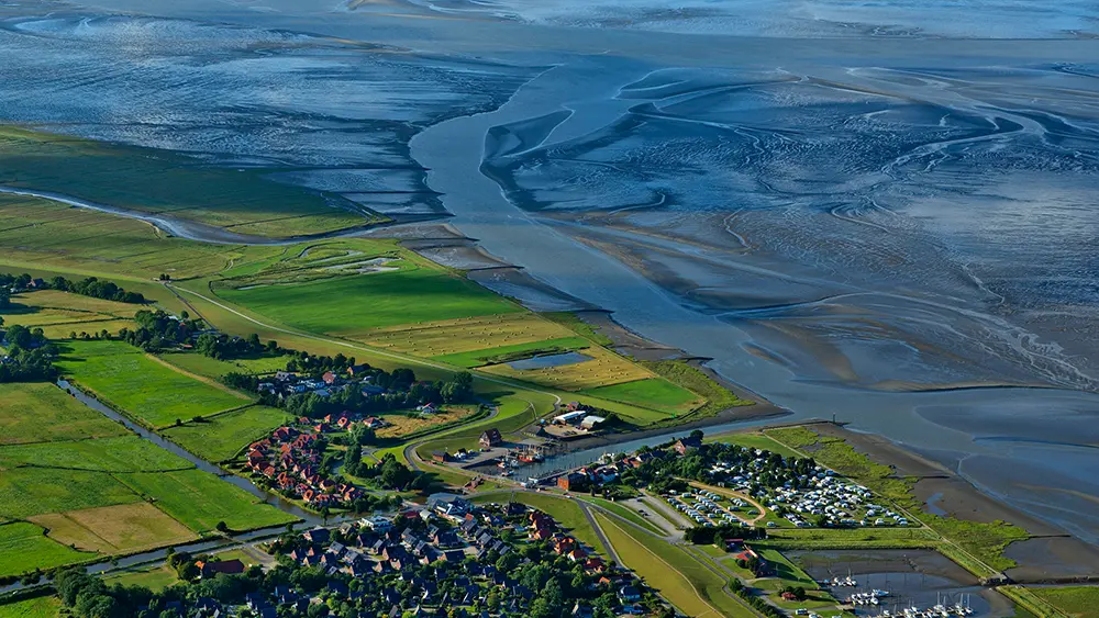 Luftaufnahme eines Küstenorts an der Nordsee mit Hafen, Campingplatz und Siedlung hinter dem Deich, davor das Wattenmeer bei Niedrigwasser mit Prielen.