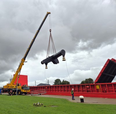 Mobilkran hebt ein großes schwarzes Stahlbauteil über die rote Klappbrücke der Nesserlander Schleuse in Emden; ein Bauarbeiter mit Warnweste und Helm beobachtet den Vorgang.