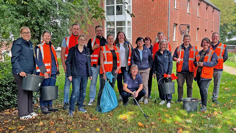 Rund 15 Mitarbeitende, teils in orangefarbenen Warnwesten, stehen mit Eimern, Greifzangen und Müllsack auf einer Wiese mit Herbstlaub vor einem Backsteingebäude.