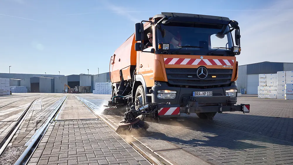 Eine orangefarbene Kehrmaschine von Niedersachsen Ports reinigt im Hafen Brake die gepflasterte Hafenfläche entlang der Gleise. Im Hintergrund sind Lagerhallen und gestapelte Ware zu sehen.