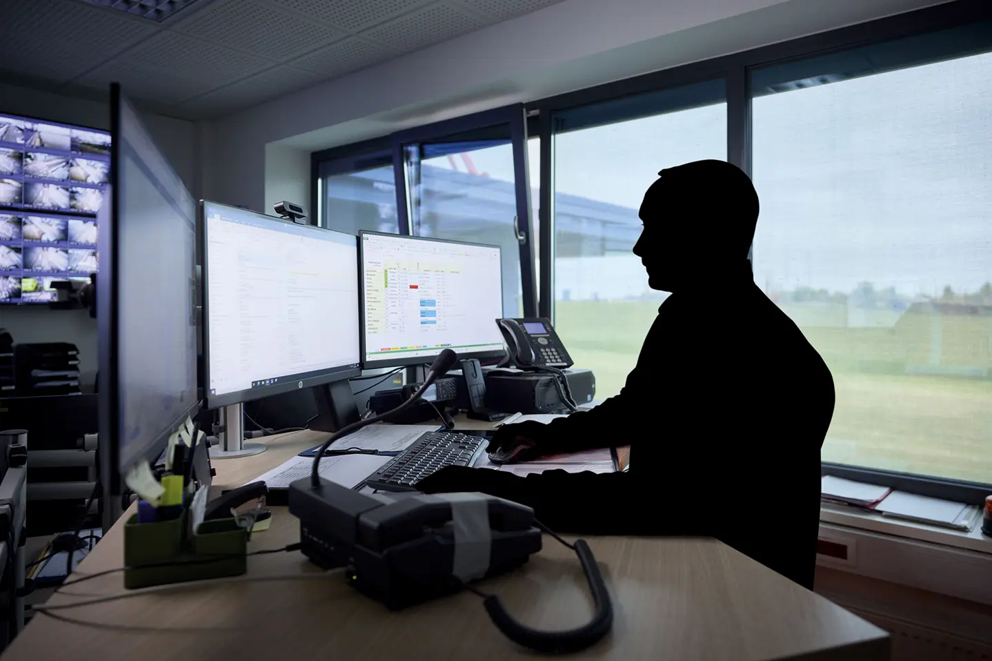 Geschwärzte Umrisse einer Person an einem Schreibtisch mit zwei Monitoren in einem abgedunkelten Büro 