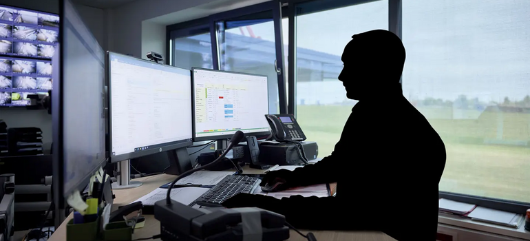 Geschwärzte Umrisse einer Person an einem Schreibtisch mit zwei Monitoren in einem abgedunkelten Büro 