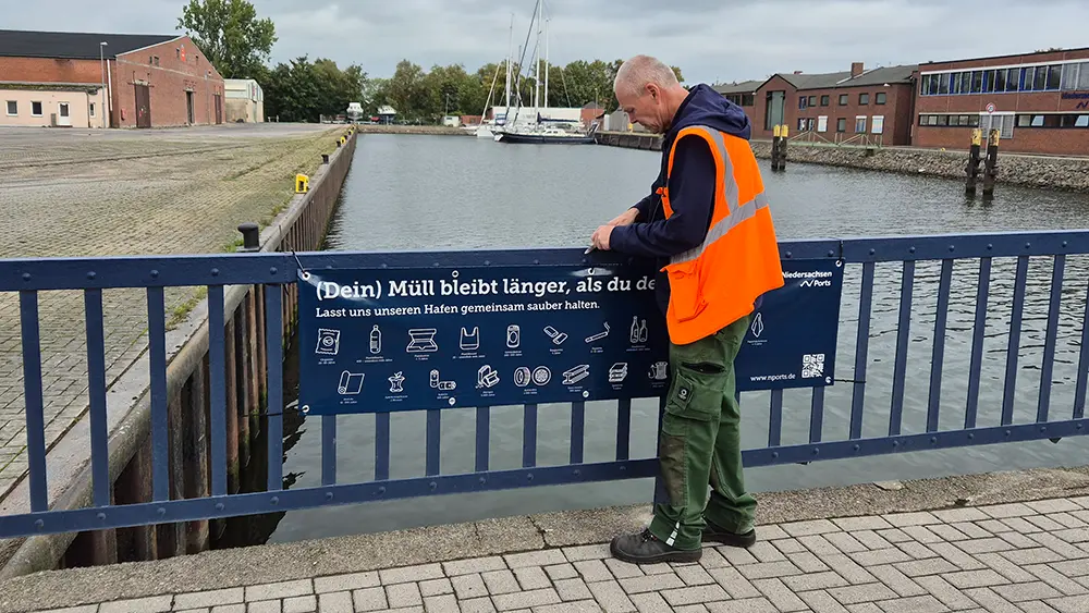 Ein Mitarbeiter von Niedersachsen Ports befestigt am Geländer eines Hafenbeckens in Brake ein Banner mit der Aufschrift „(Dein) Müll bleibt länger, als du denkst. Lasst uns unseren Hafen gemeinsam sauber halten.“