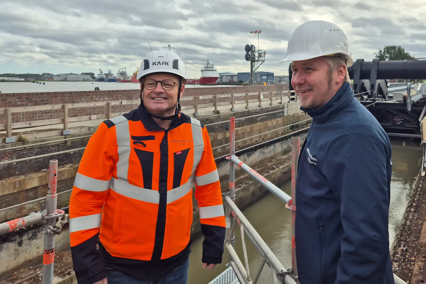 NPorts-Projektleiter Wilfried Schleusener (links, orange Warnjacke) und Niederlassungsleiter Aiko Hollander (rechts) auf einem Gerüst an der Großen Seeschleuse Emden, dahinter Hafen und Schiffe.