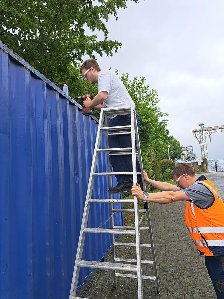 Ein Mitarbeiter bohrt auf einer Leiter am oberen Rand eines blauen Containers, ein Kollege in Warnweste sichert die Leiter. Im Hintergrund sind Hafenanlagen zu sehen.