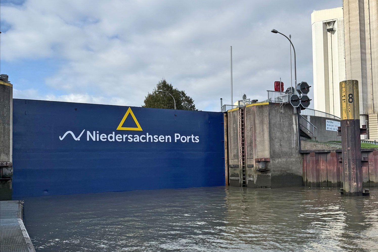 Geschlossenes, blau lackiertes Außenhaupttor der Schleuse im Hafen Brake mit dem Schriftzug und Logo von Niedersachsen Ports. Im Vordergrund die gefüllte Schleusenkammer mit Steganlage, rechts die Schleusenmauer mit Signalanlage und dahinter hohe Silogebäude.
