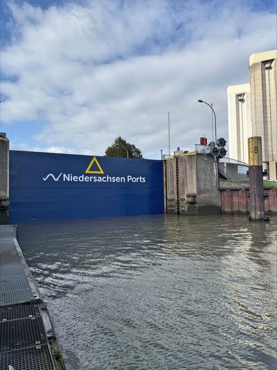 Geschlossenes, blau lackiertes Außenhaupttor der Schleuse im Hafen Brake mit dem Schriftzug und Logo von Niedersachsen Ports. Im Vordergrund die gefüllte Schleusenkammer mit Steganlage, rechts die Schleusenmauer mit Signalanlage und dahinter hohe Silogebäude.