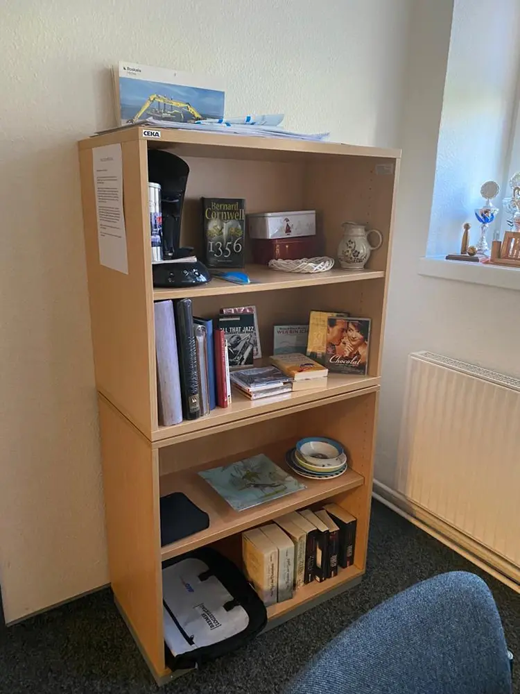 Ein helles Holzregal mit gespendeten Sachspenden: Bücher, DVDs, eine Kaffeemaschine, Geschirr, eine Tasche und kleine Dekoartikel.