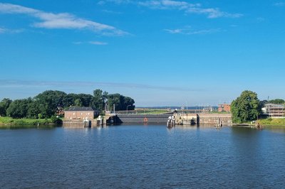 Nesserlander Schleuse in Emden bei blauem Himmel, vom Wasser aus gesehen: geschlossenes Schleusentor mit Dalben, dahinter das Backsteingebäude der Schleusenanlage und die Ems.