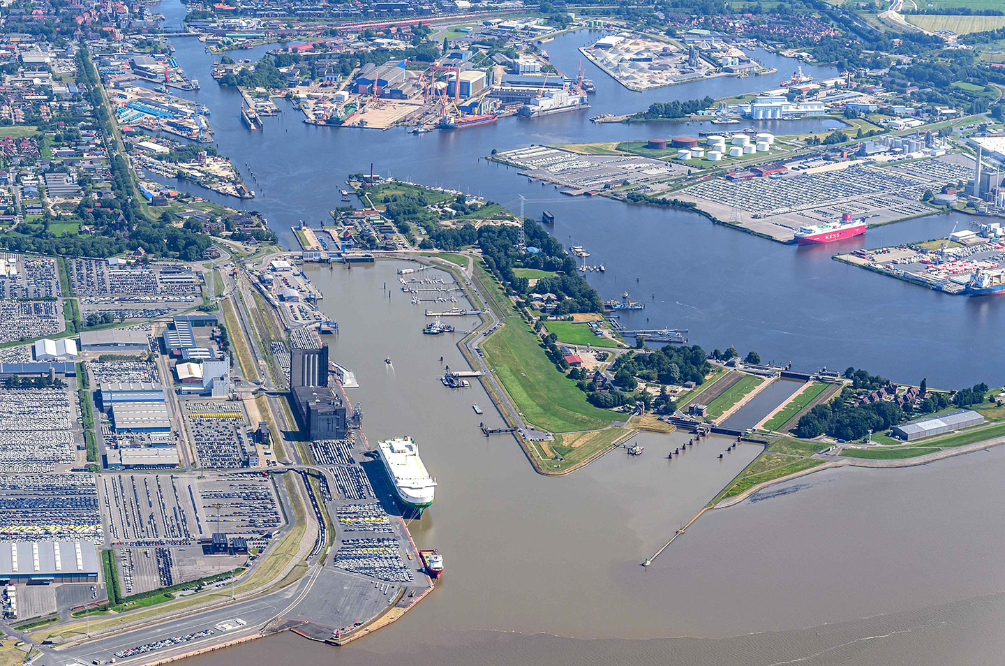 Luftbild des Hafens Emden: Große Seeschleuse mit dem Verbindungskanal zur Ems, daneben Autoterminal mit Fahrzeugstellflächen und ein Autotransporter am Kai.