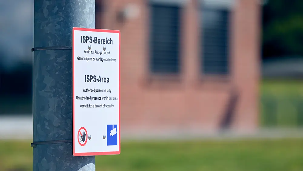 Schild an Pfosten mit Aufschrift Achtung - ISPS-Bereich, Zutritt nur mit Genehmigung. Im Hintergrund unscharf umrisse eines Gebäudes
