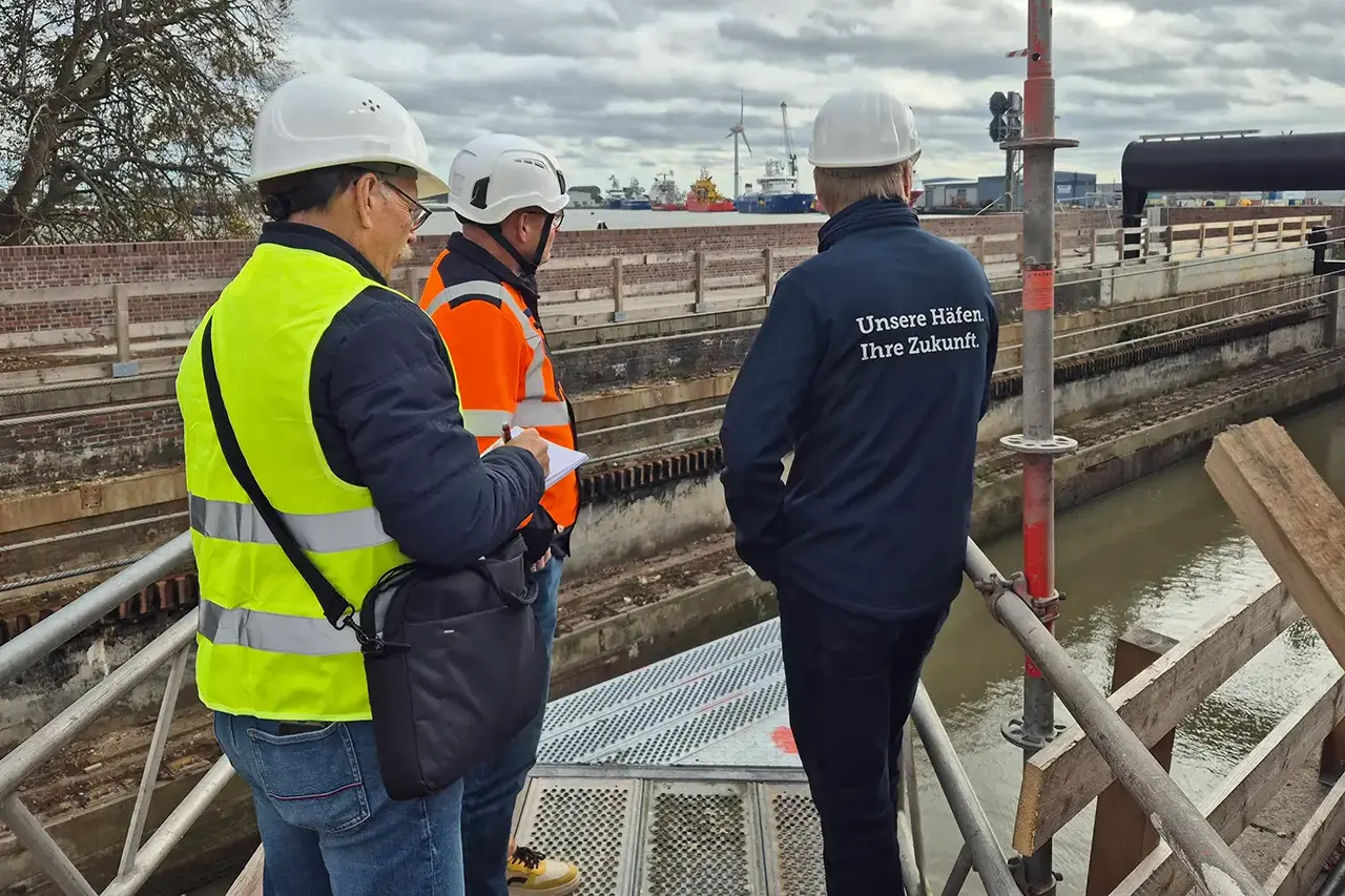 Begehung auf dem Gerüst Drei Personen mit Schutzhelmen stehen auf einer Gerüstplattform und blicken in die Schleusenanlage. Im Hintergrund liegen Schiffe im Emder Hafen.